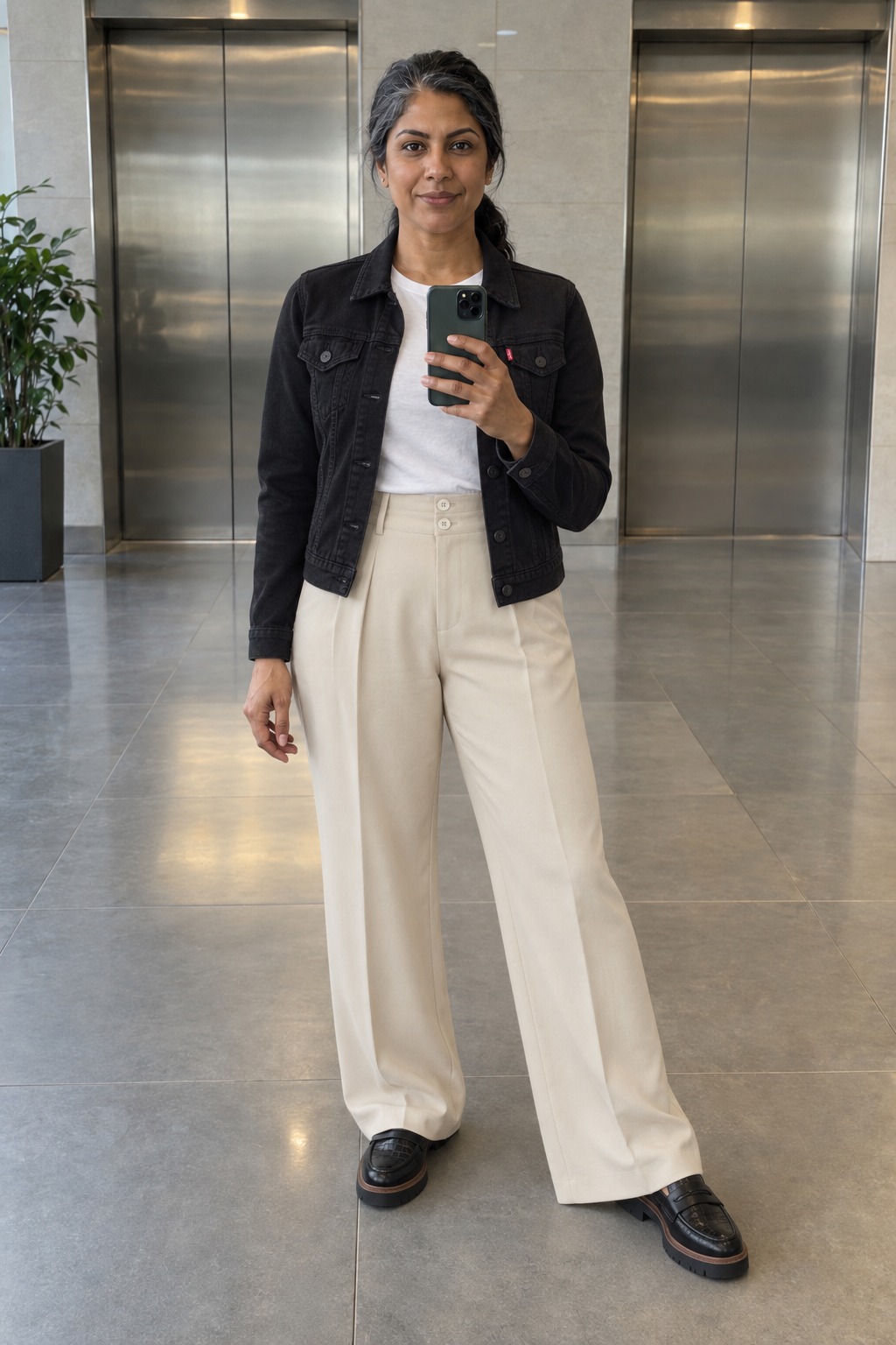 Woman taking a full-length mirror selfie by the office lifts wearing a black Levi's denim trucker jacket over a white tee, high-waisted cream wide-leg trousers and black lug-sole Clarks loafers.