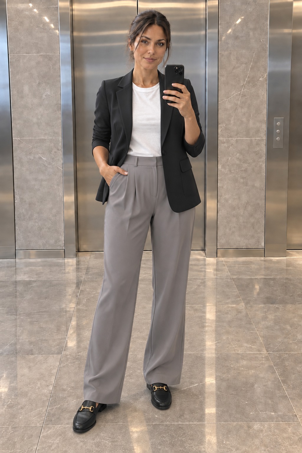 Woman taking a full-length mirror selfie in an office lobby wearing an open black single-button blazer over a white tee, straight-leg grey tailored trousers and black leather loafers.