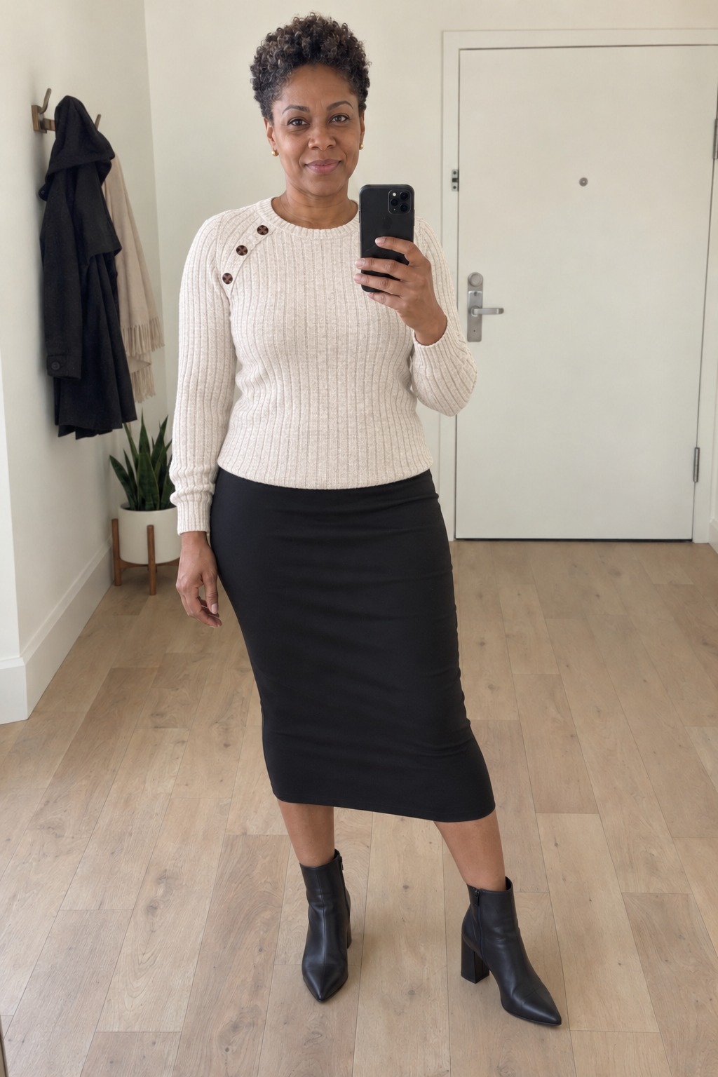 Woman taking a full-length mirror selfie in an apartment entrance hall wearing a cream ribbed-knit sweater over a black pencil skirt with black pointed block-heel ankle boots.