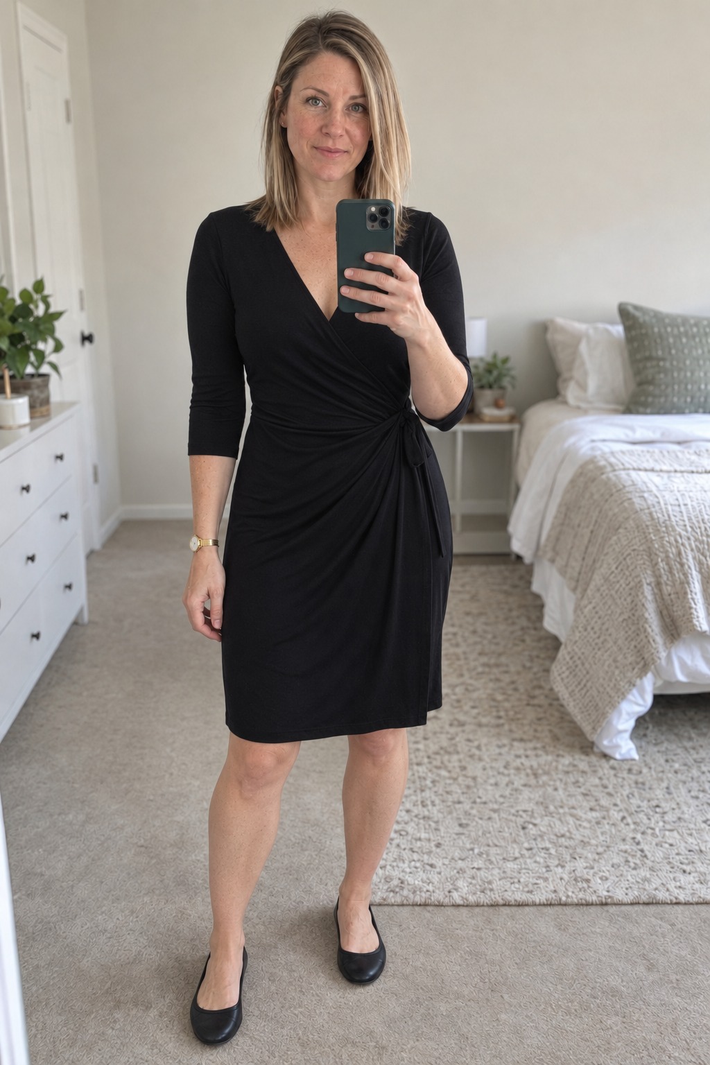 Woman taking a full-length mirror selfie in a bedroom wardrobe mirror wearing a black knee-length wrap dress with three-quarter sleeves and black ballet flats.
