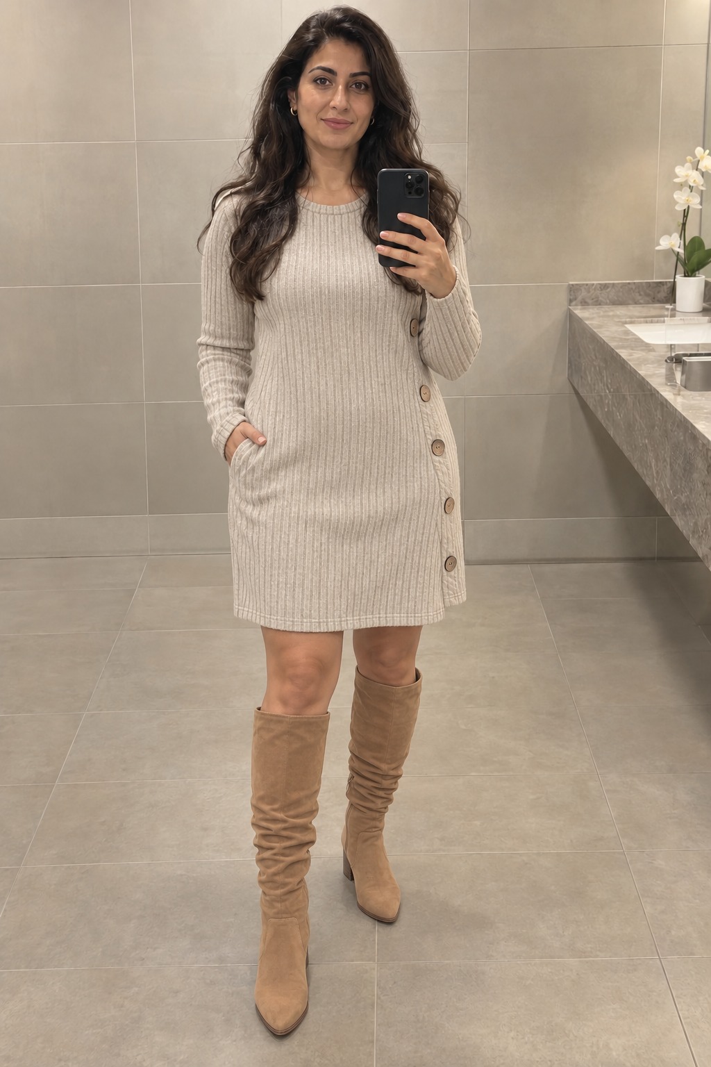 Woman taking a full-length mirror selfie in an office washroom wearing an oatmeal ribbed sweater dress with side buttons and tan suede slouchy knee-high boots.