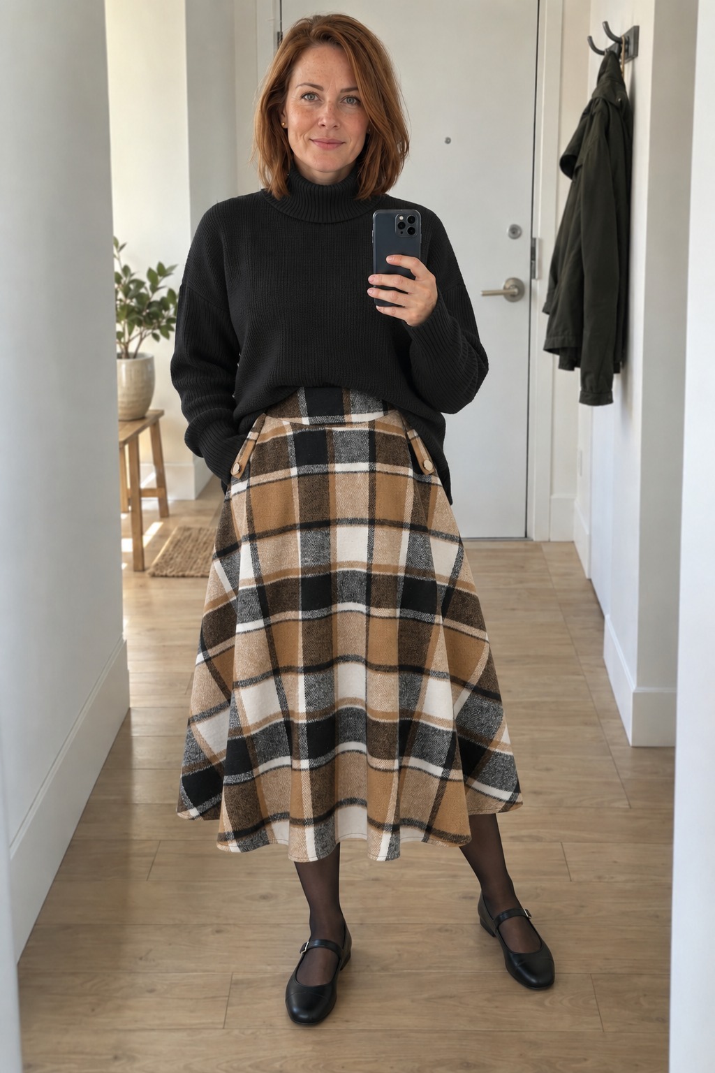 Woman taking a full-length mirror selfie in an apartment entrance hall wearing a black turtleneck tucked into a camel, black and cream plaid midi skirt with sheer black tights and black Mary Jane flats.