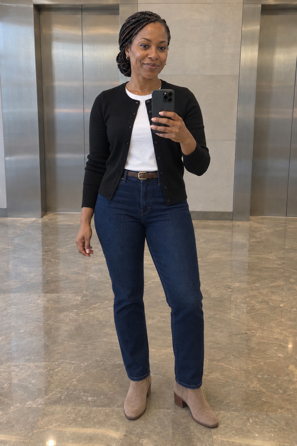 Woman taking a full-length mirror selfie by the office lifts wearing a black cardigan over a white tee tucked into dark straight jeans with a brown belt and taupe suede ankle boots.
