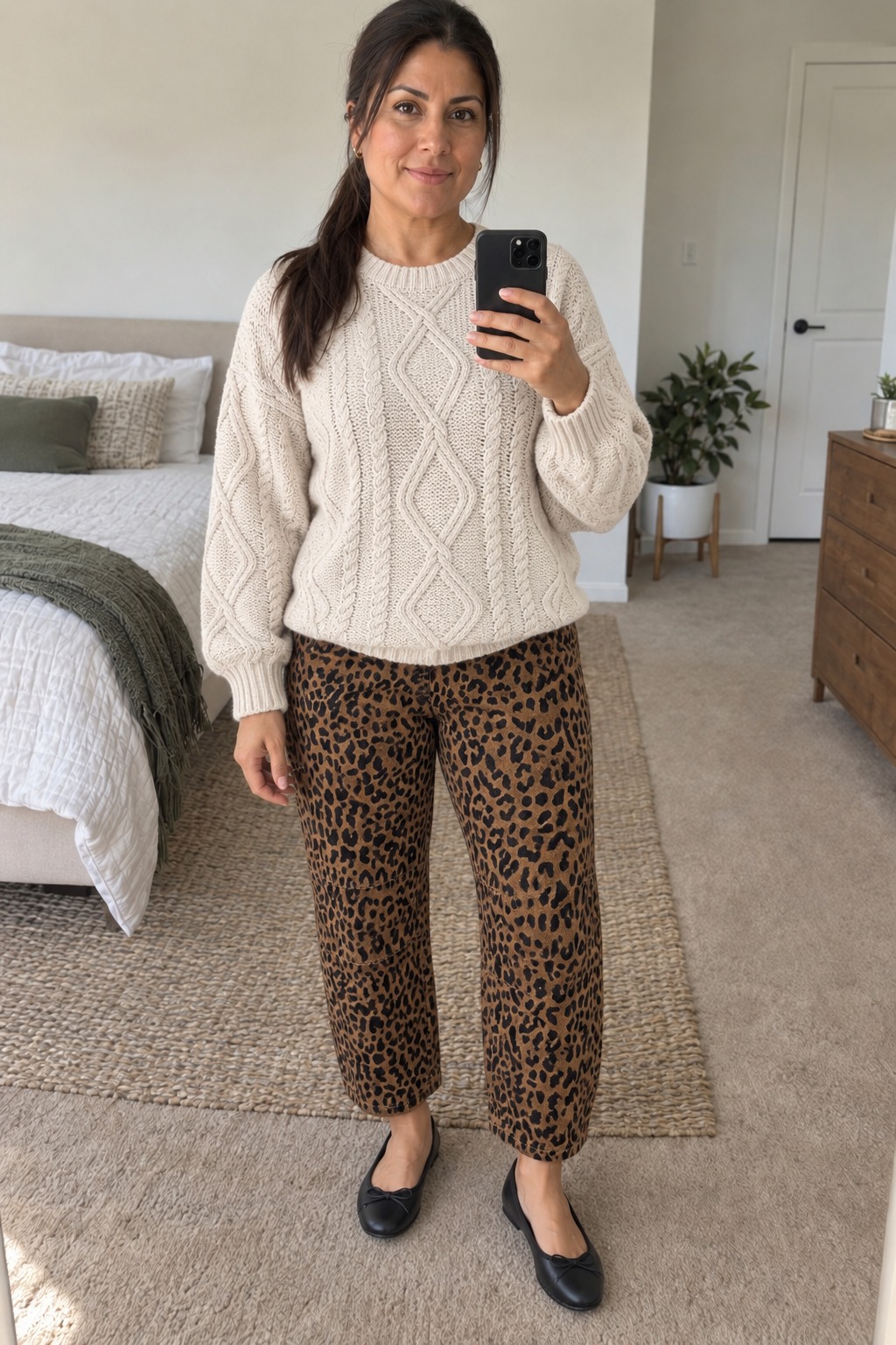 Woman taking a full-length mirror selfie in a bedroom wearing a cream oversized cable-knit sweater over leopard-print trousers with black bow ballet flats.
