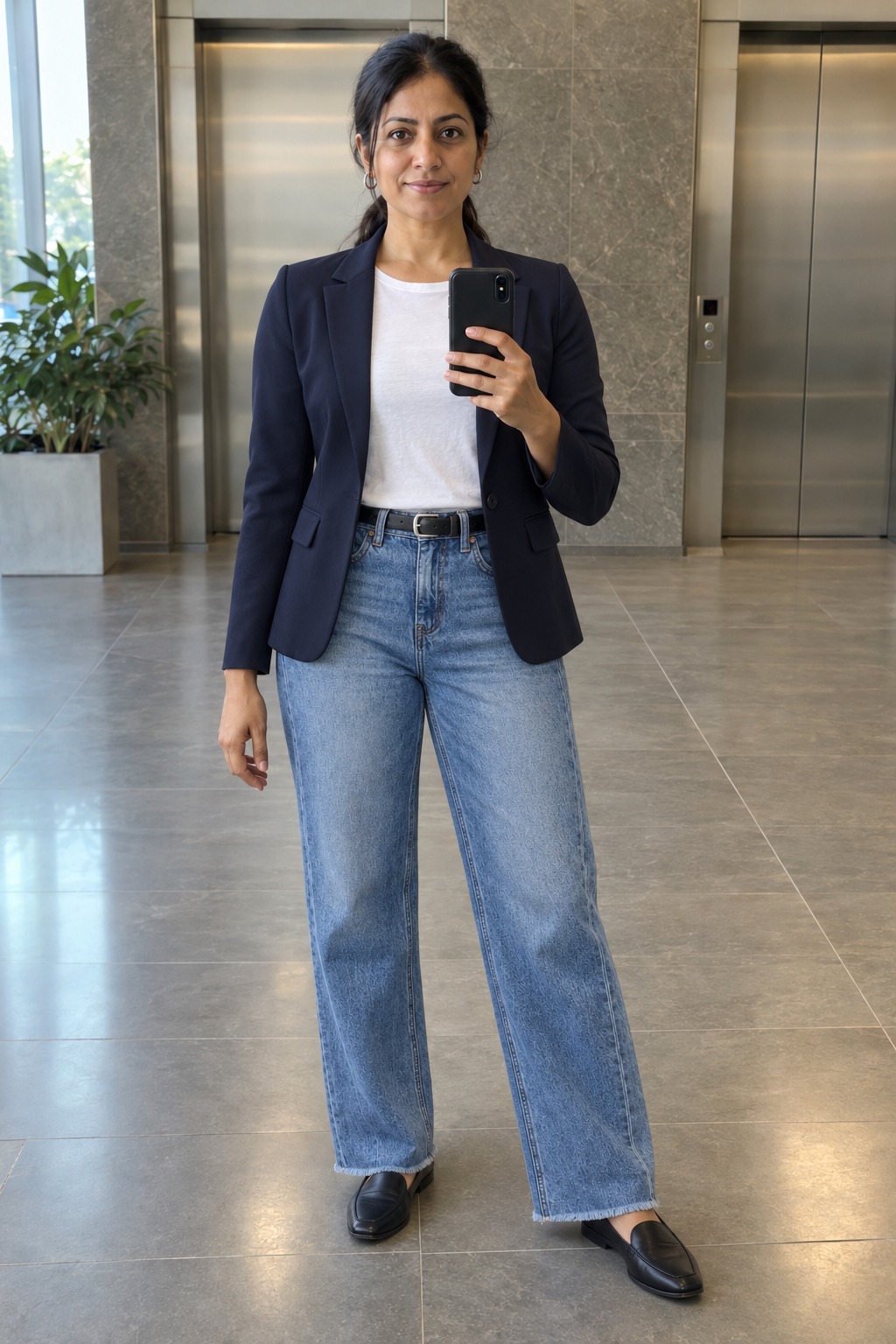 Woman taking a full-length mirror selfie by the office elevators wearing an open navy blazer over a white tee tucked into medium-blue wide-leg jeans, a slim black belt, small silver hoops and black loafers.