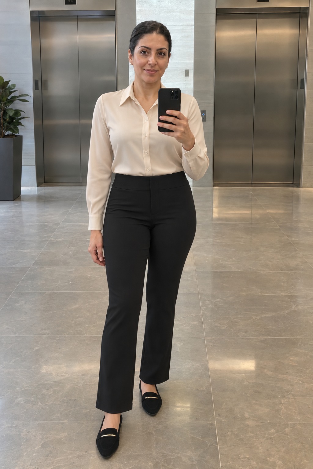 Woman taking a full-length mirror selfie by the office elevators wearing a cream long-sleeve blouse tucked into black trousers with black pointed-toe flats, gold-tone shoe accents and small pearl studs.