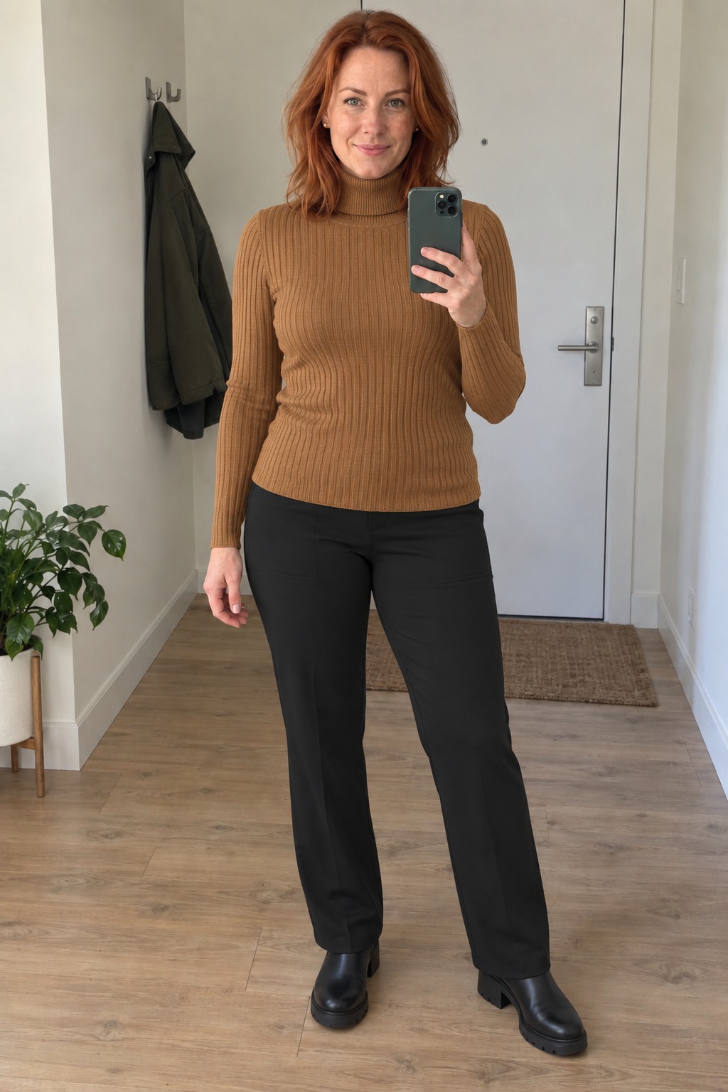 Woman taking a full-length mirror selfie in an entryway with a white door, wood floor and potted plant, wearing a camel ribbed turtleneck untucked over black trousers and chunky black Chelsea boots with small gold stud earrings.