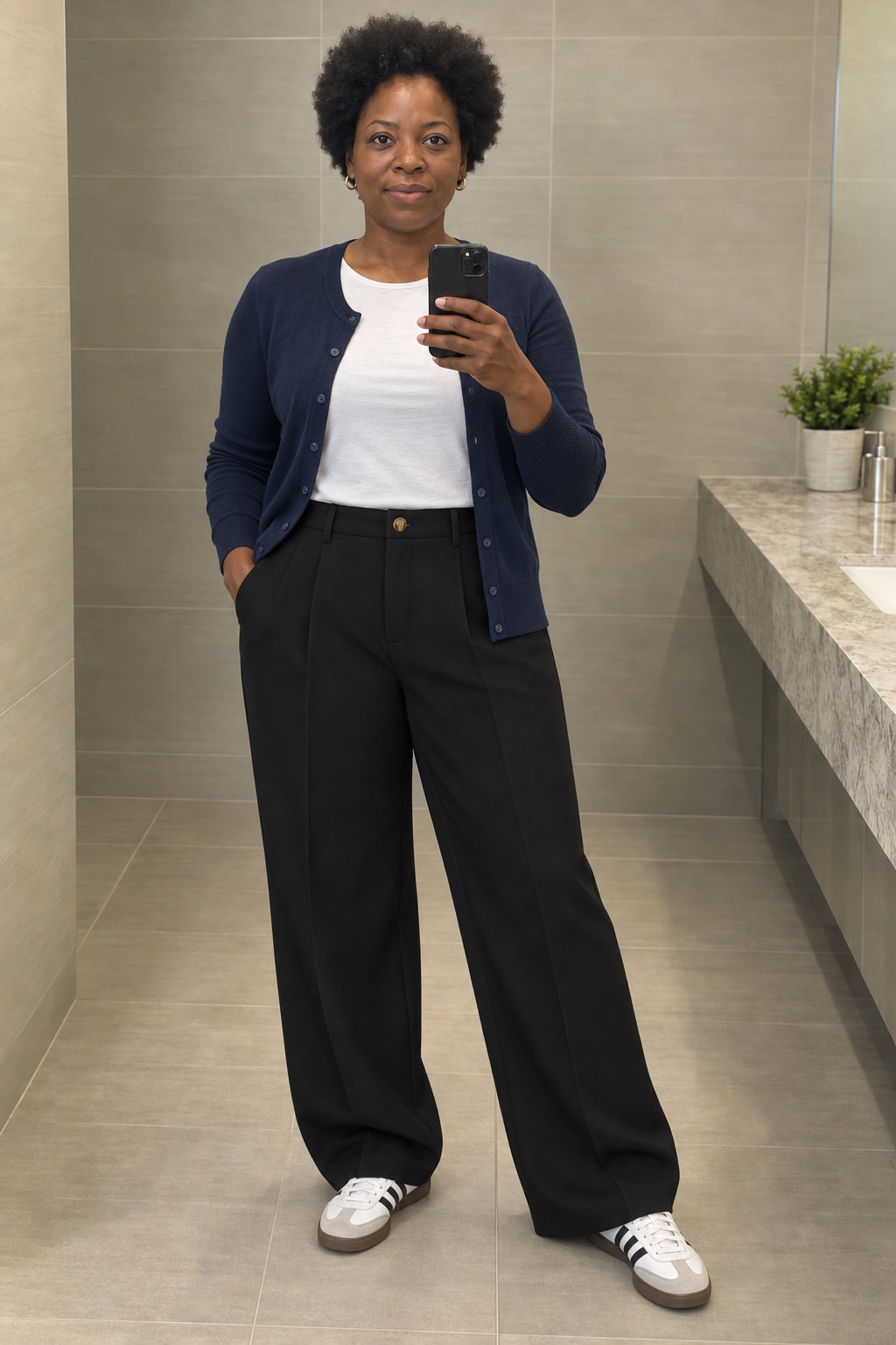 Woman taking a full-length mirror selfie in a tiled restroom beside a stone vanity, wearing an open navy cardigan over a tucked white tee, black wide-leg trousers, white sneakers with black stripes and small gold hoops.