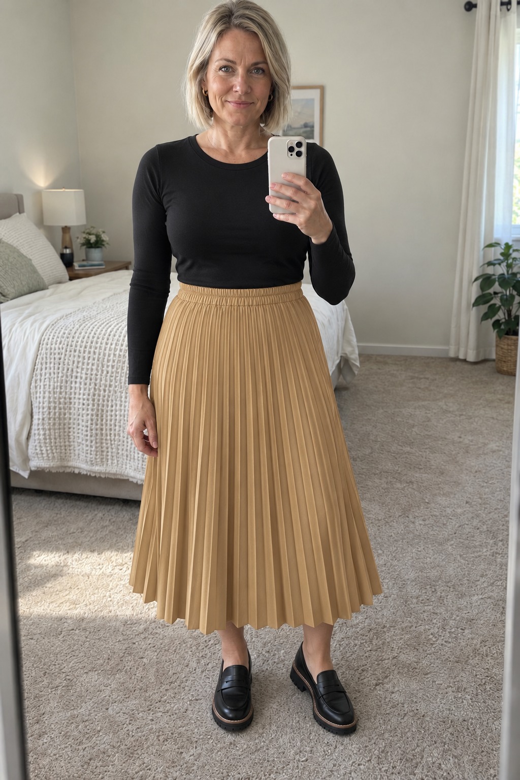 Woman taking a full-length mirror selfie in a neutral bedroom wearing a black fitted long-sleeve crew-neck top tucked into a camel pleated midi skirt, black chunky penny loafers and small gold hoops.