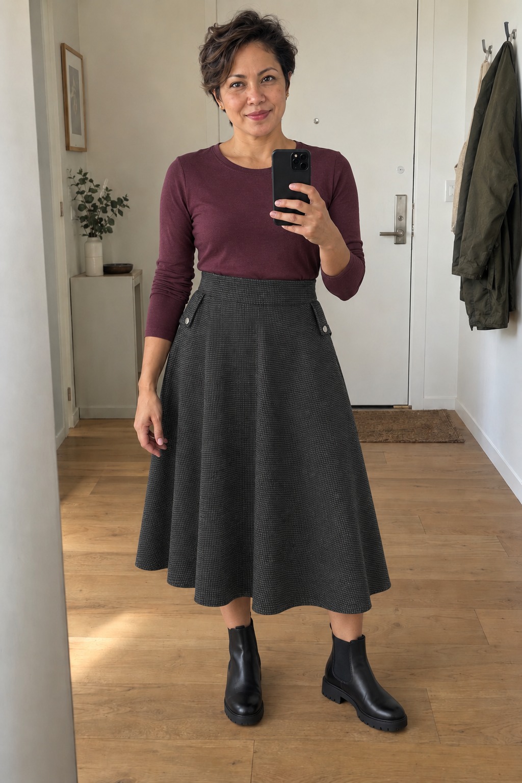 Woman taking a full-length mirror selfie in a home entryway with a wood floor, wearing a burgundy long-sleeve top tucked into a charcoal patterned A-line midi skirt with black lug-sole Chelsea boots and small gold stud earrings.