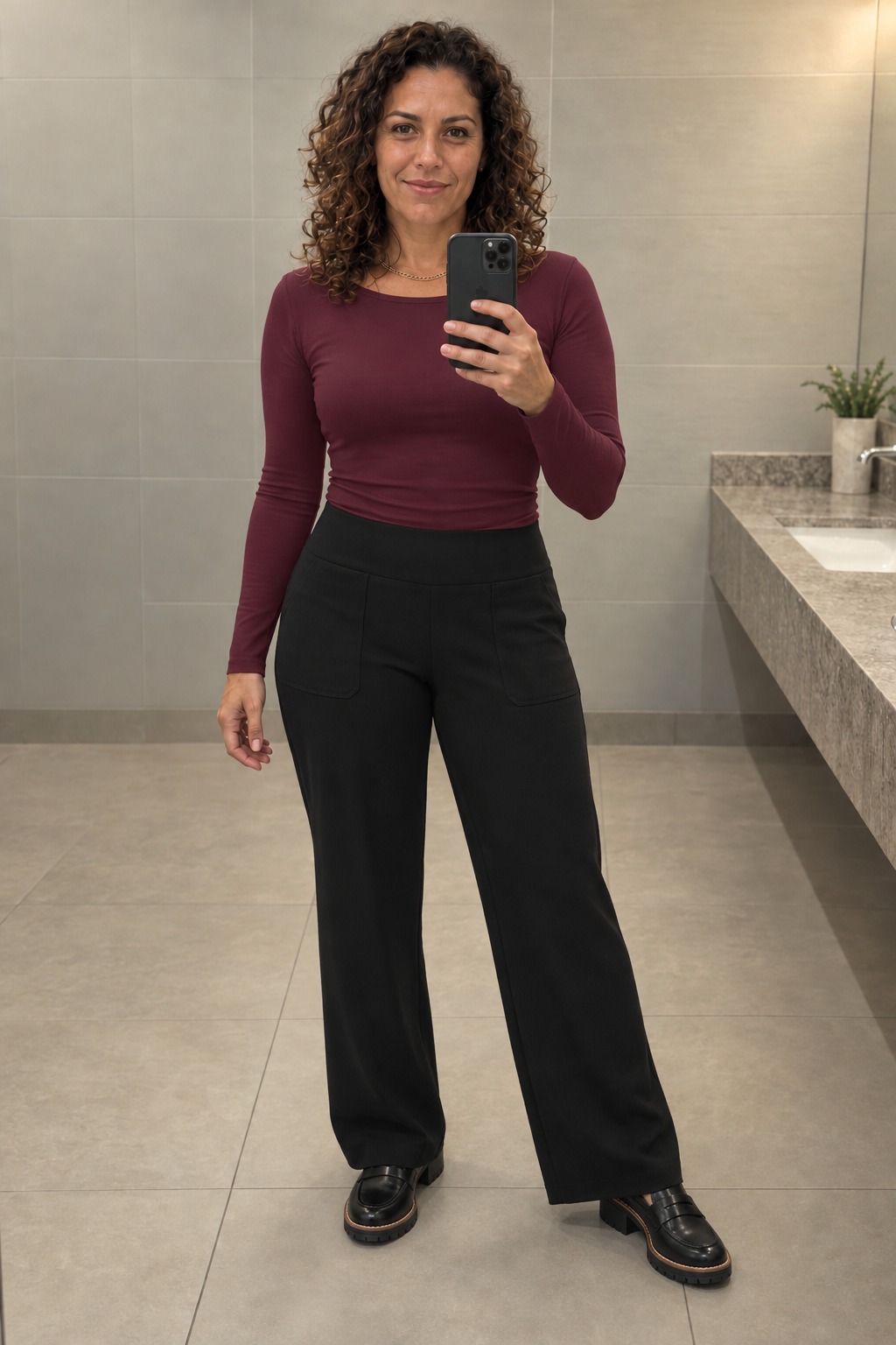 Woman taking a full-length mirror selfie in a beige-tiled restroom wearing a fitted burgundy long-sleeve top tucked into black wide-leg trousers, black chunky penny loafers and a thin gold necklace.