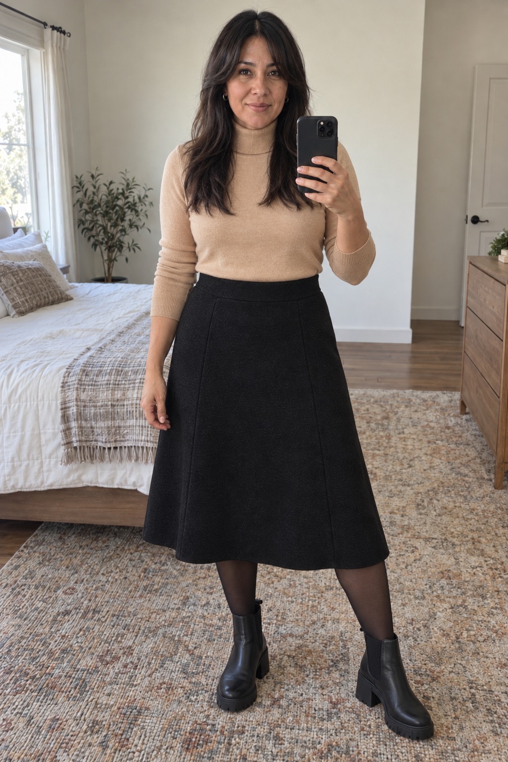 Woman taking a full-length mirror selfie in a bedroom wearing a camel turtleneck tucked into a black A-line midi skirt, sheer black tights, black chunky Chelsea boots and small silver hoops.