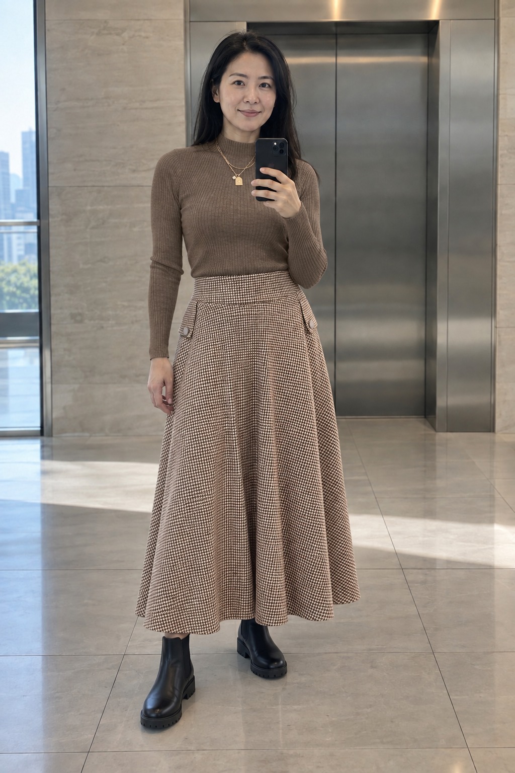 Woman taking a full-length mirror selfie by office elevators wearing a mocha ribbed mock-neck sweater tucked into a brown-and-cream patterned maxi skirt, a small gold pendant necklace and black Chelsea boots.
