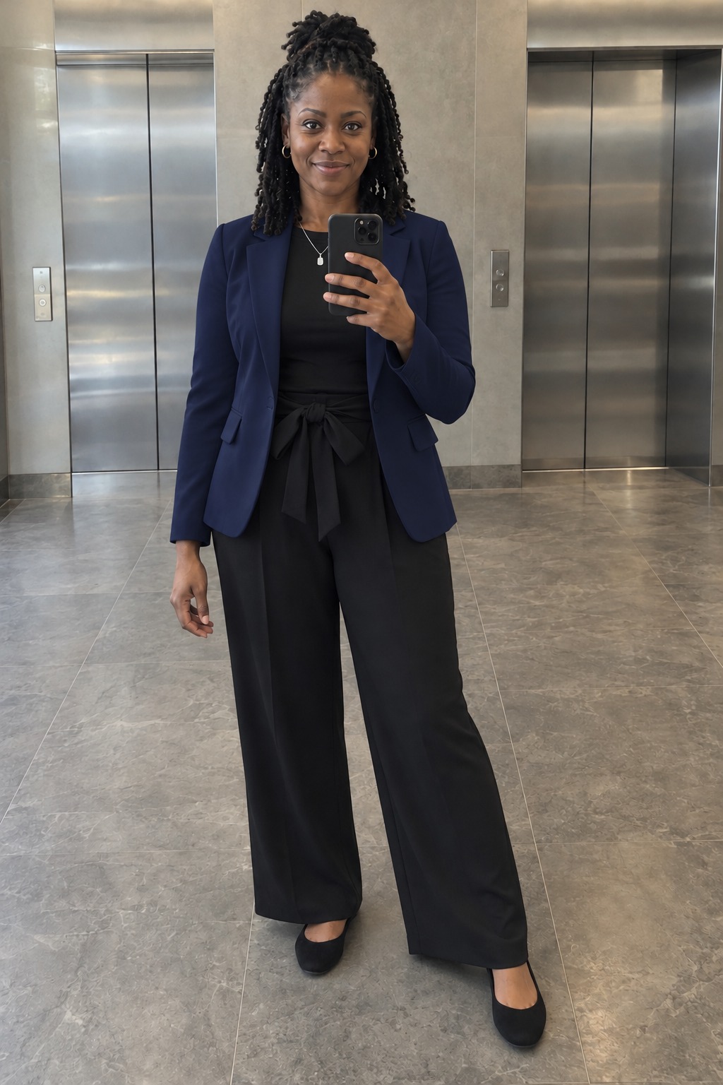 Woman taking a full-length mirror selfie by office elevators wearing an open navy blazer over a tucked black top, black wide-leg tie-waist trousers, black round-toe ballet flats and a thin silver pendant necklace.