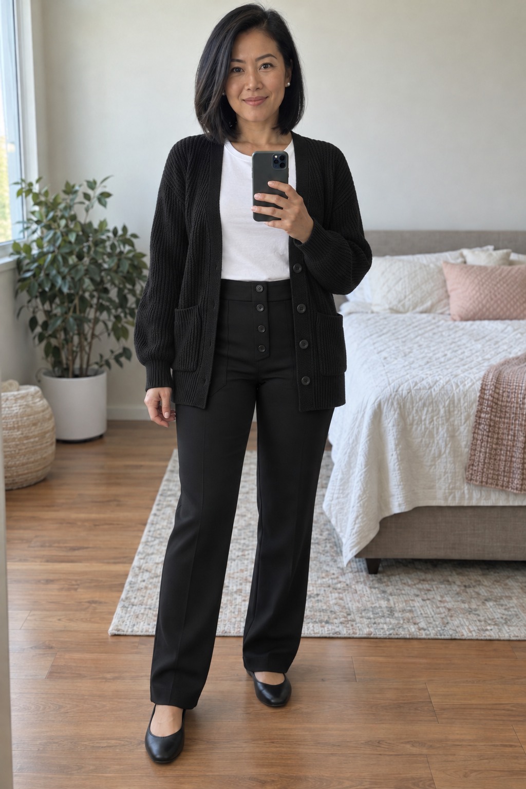 Woman taking a full-length mirror selfie in a bright bedroom wearing an open black chunky cardigan over a white tee tucked into black button-front trousers, with black ballet flats and small pearl studs.