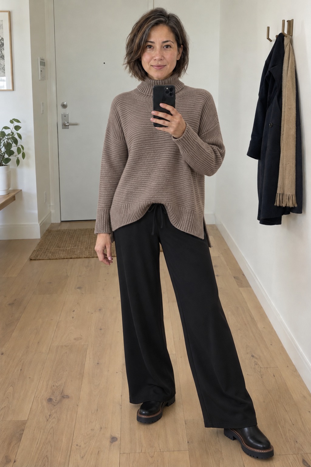 Woman taking a full-length mirror selfie in a bright entryway with wood flooring, wearing a loose taupe ribbed roll-neck sweater, black wide-leg pants and black lug-sole Chelsea boots, with a coat and scarf hanging behind her.